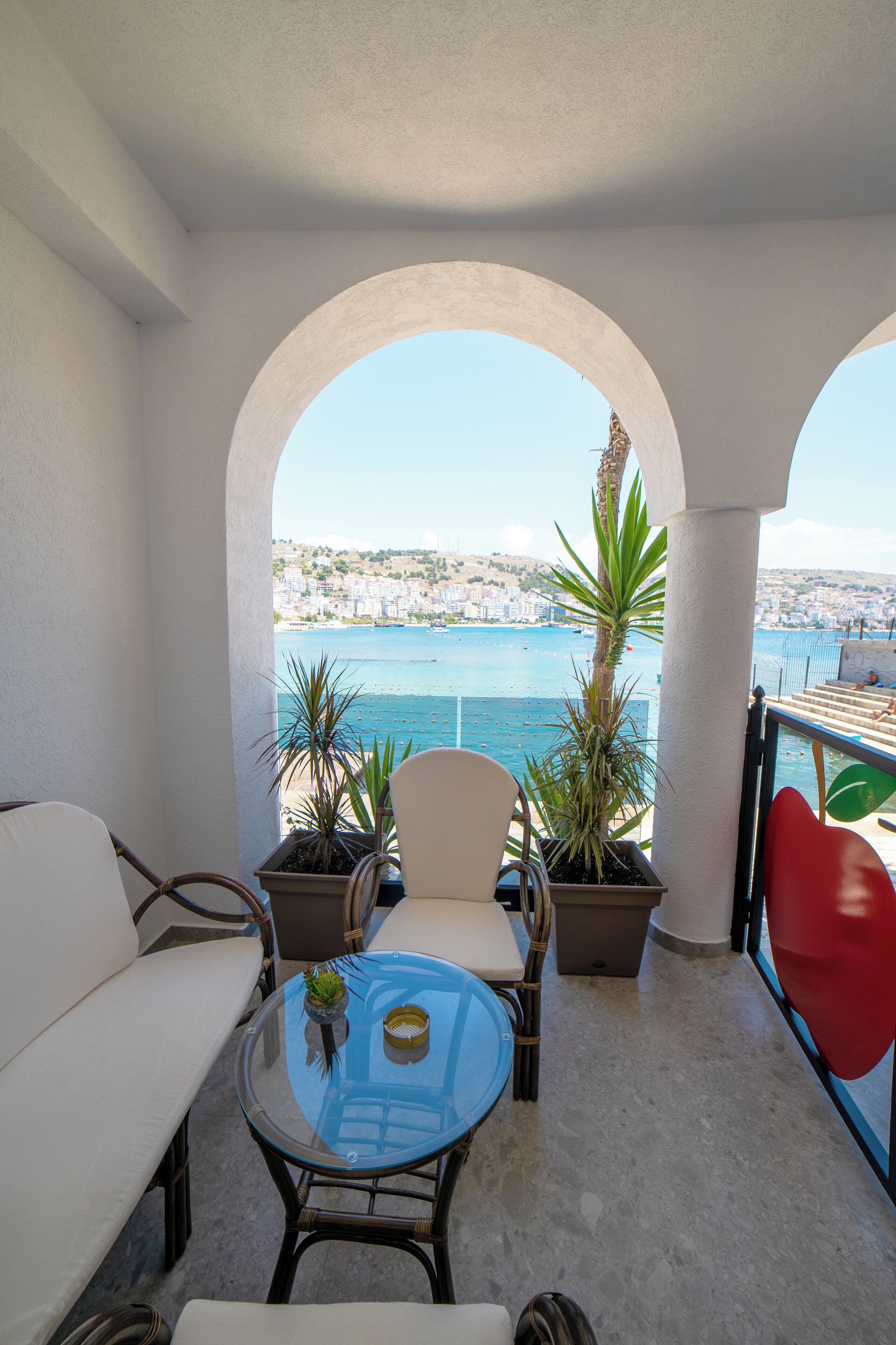 A balcony with a glass table, two chairs, a sofa, and potted plants.