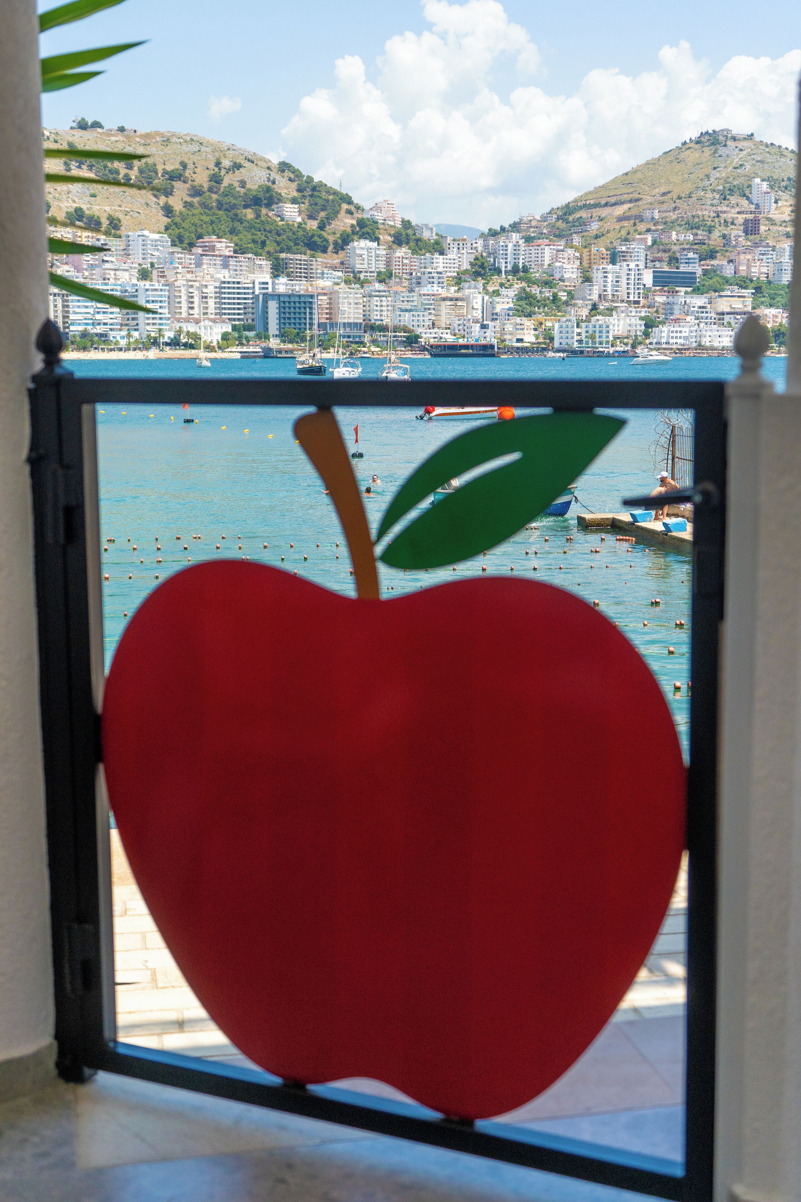 Una decorazione a forma di mela rossa su una ringhiera di balcone con vista su una città costiera, edifici e colline.