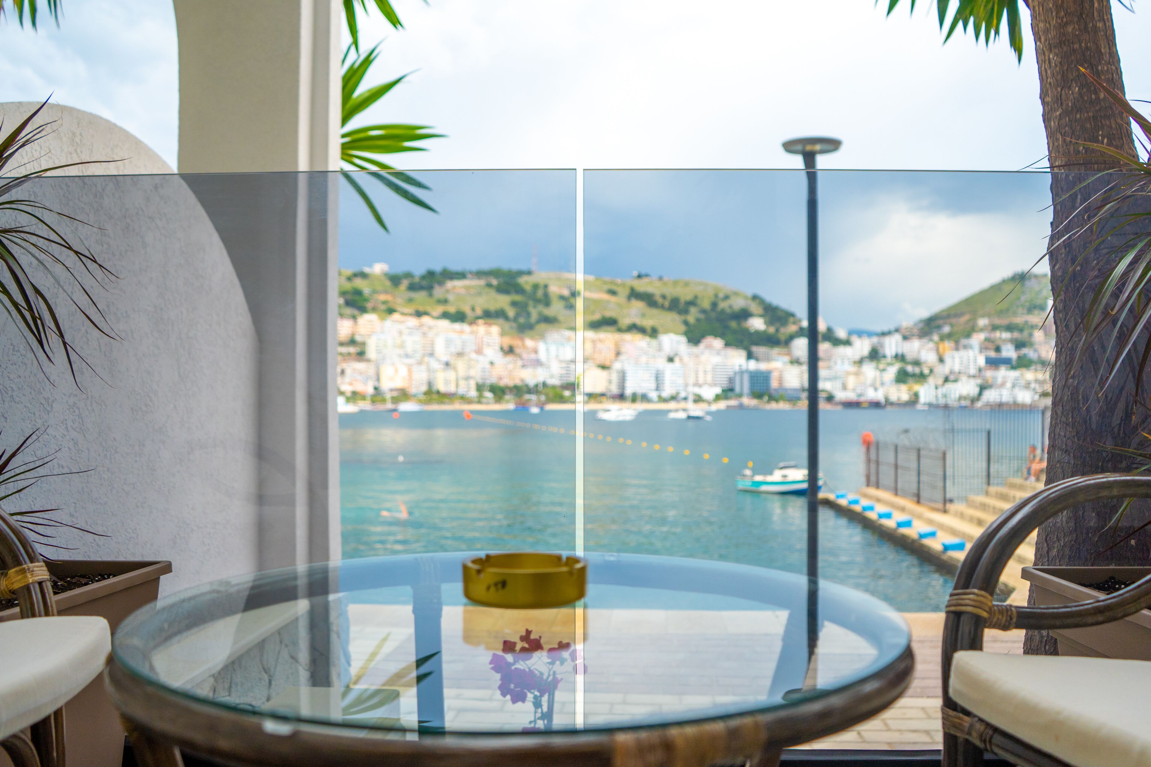 A balcony with a glass table, chairs, and a view of a coastal cityscape with buildings and boats.