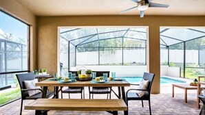 Un espace patio aménagé avec une table et des chaises en bois, un banc et une structure en verre offrant une vue sur une piscine et sur la verdure.