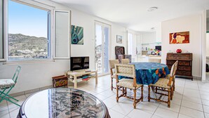 A spacious living area with a dining table, chairs, a glass-top coffee table, a TV, and a view of a hillside.