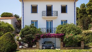 A two-story building with a car parked in front, surrounded by lush greenery and flowering plants.