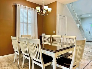 Un coin repas avec une table en bois et des chaises blanches, un lustre et un escalier en arrière-plan.