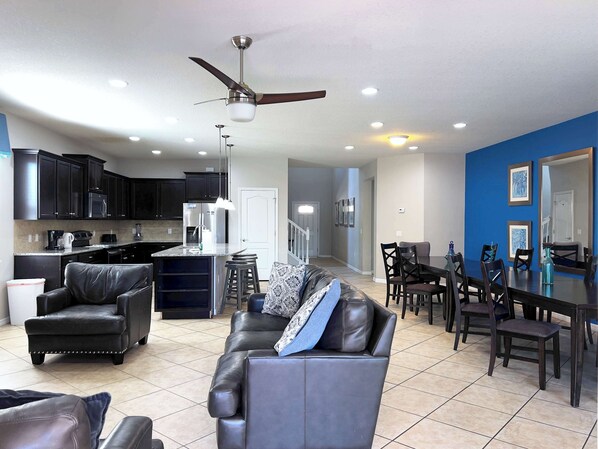 A spacious living room with a leather sofa, a dining area with a table and chairs, a kitchen with dark cabinets and modern appliances, and a ceiling fan.