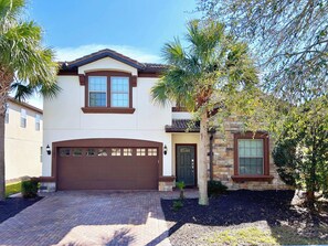 A two-story house with a brown garage door, a stone exterior, and a small front yard with palm trees and shrubs.