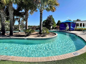 A clear swimming pool with a wooden deck edge, surrounded by palm trees and a covered seating area.
