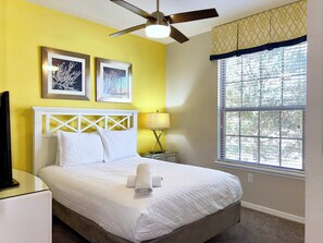 A neatly made bed with white bedding and a rolled towel, a ceiling fan, two framed pictures on the wall, a bedside lamp, and a window with blinds.