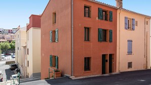 A two-story building with a red facade and green shutters, featuring a small balcony and a visible entrance.