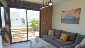 A modern living room with a grey sofa, a wooden coffee table, and a sliding glass door leading to an outdoor patio with wooden benches.