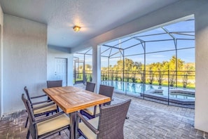 Une terrasse couverte, avec une table en bois et des chaises en osier, donnant sur une piscine et un jardin.