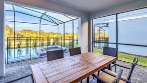 A modern outdoor dining area with a wooden table and wicker chairs, overlooking a swimming pool and a landscaped garden. - Big Screen-Gameroom-Private Pool-6mi 2 Disney-8911 (Four Corners)