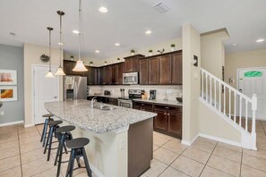A kitchen with a granite island, bar stools, dark wooden cabinets, stainless steel appliances, and pendant lighting.