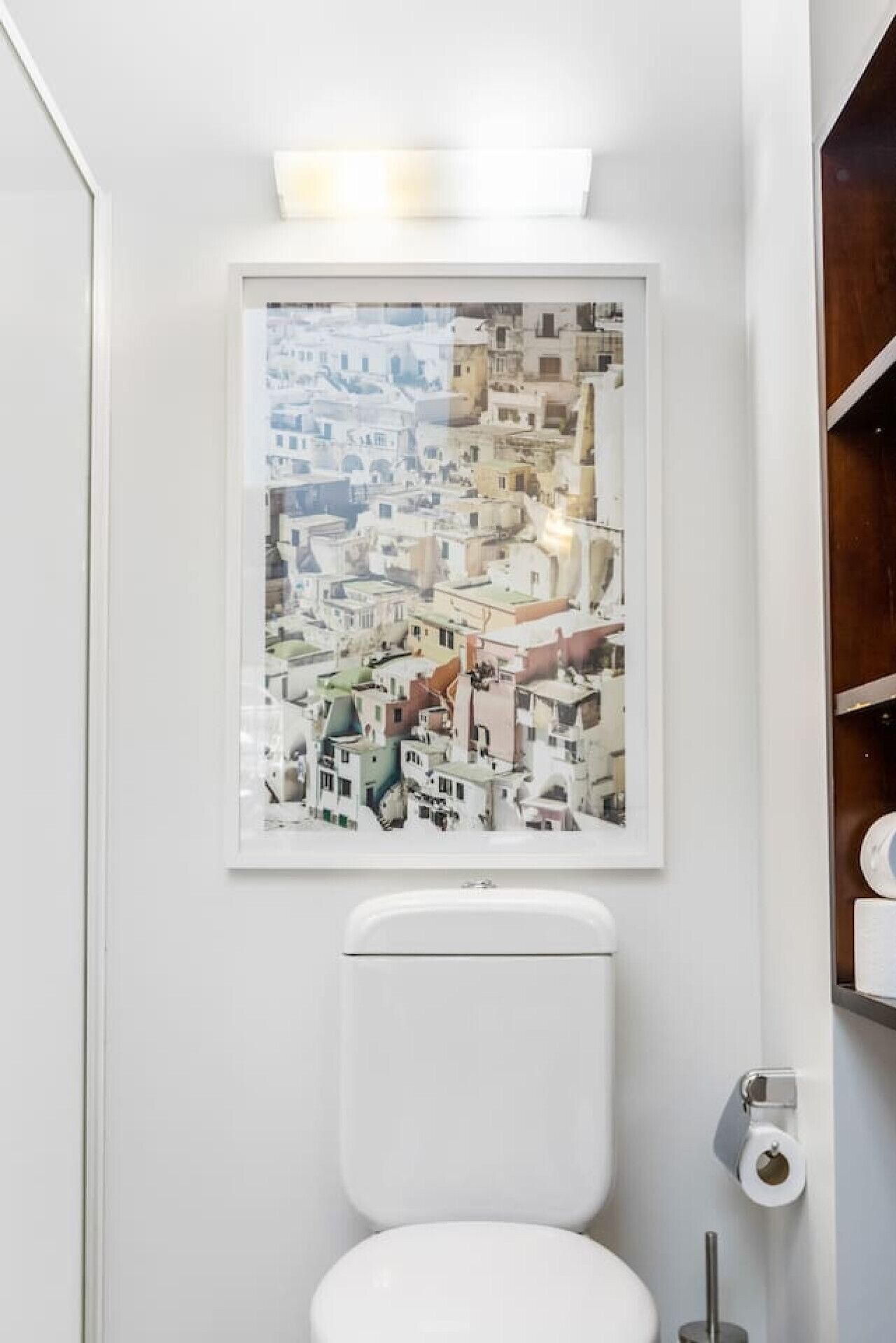 Un baño con un inodoro blanco, una fotografía enmarcada de un paisaje urbano en la pared y un estante de madera con artículos de aseo.