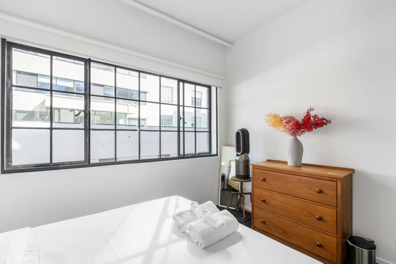 A hotel room with a large window, a bed with white linens, a wooden dresser, and a vase with flowers.