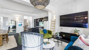 A modern living room with a large flat-screen TV, a dining area with a wooden table and chairs, and a kitchen in the background. - Gorgeous NY Style Loft in CBD | Netflix Wifi | (Auckland CBD)