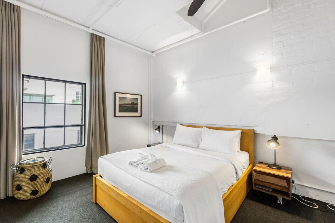 Une chambre d’hôtel avec une grande fenêtre, un lit avec du linge de maison blanc, une lampe de chevet et une petite table de nuit en bois.