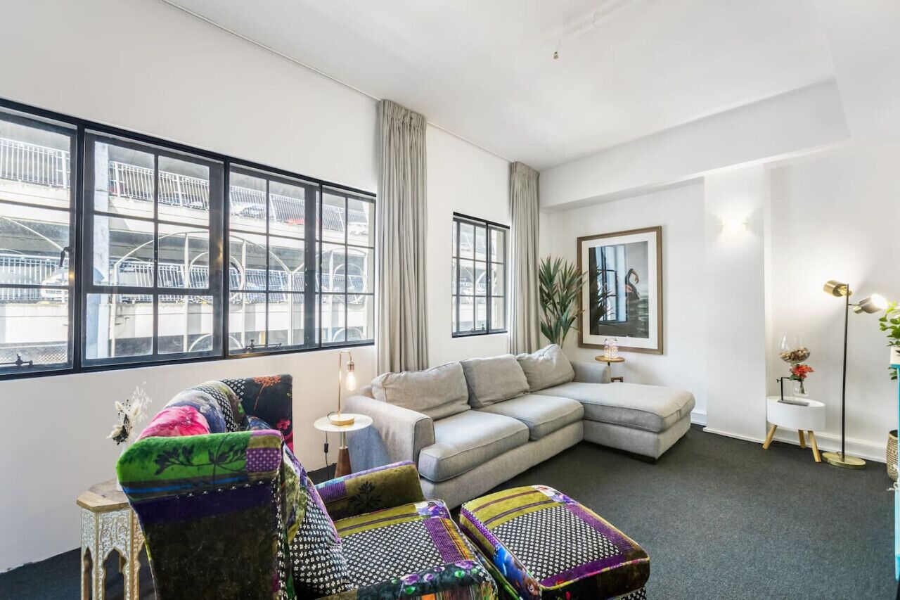 Un salon moderne avec un canapé d’angle gris, des fauteuils colorés, une petite table d’appoint et une grande fenêtre donnant sur un parking.