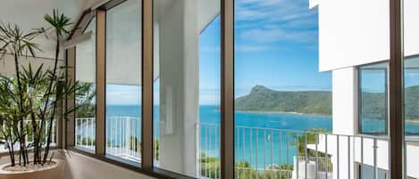 A balcony with a view of the sea and a mountain, a potted plant, and a white railing.