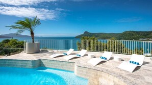 Un espace aménagé au bord de la piscine, avec des chaises longues et un palmier, offrant une vue sur une étendue d’eau et des collines.
