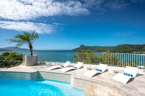 Un espace aménagé au bord de la piscine, avec des chaises longues et un palmier, offrant une vue sur une étendue d’eau et des collines.