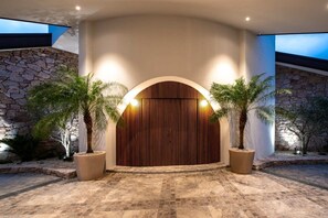 A modern entrance with a stone wall, a wooden door, and two potted palm trees. - Casa Rotunda (Whitsundays)