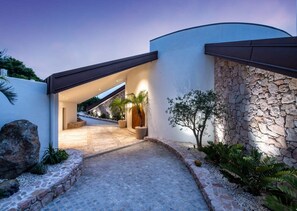 A modern building with a curved facade, a stone wall, and a paved entrance area with plants. - Casa Rotunda (Whitsundays)