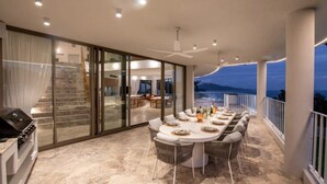 Un espace de repas extérieur moderne, doté d’une cuisine fermée par des parois de verre, d’une table à manger pouvant accueillir six personnes et offrant une vue sur les montagnes au crépuscule.