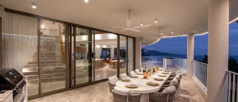 A modern outdoor dining area with a glass-enclosed kitchen, a dining table set for six, and a view of the mountains at dusk.
