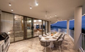 Un espace de repas extérieur moderne, doté d’une cuisine fermée par des parois de verre, d’une table à manger pouvant accueillir six personnes et offrant une vue sur les montagnes au crépuscule.