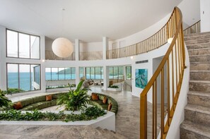 A spacious interior with a curved staircase, large windows offering ocean views, and a central seating area with plants. - Casa Rotunda (Whitsundays)