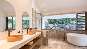 Une salle de bain moderne avec une grande baignoire, deux lavabos et une vue sur les rochers par une fenêtre.
