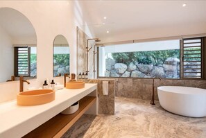 Une salle de bain moderne avec une grande baignoire, deux lavabos et une vue sur les rochers par une fenêtre.