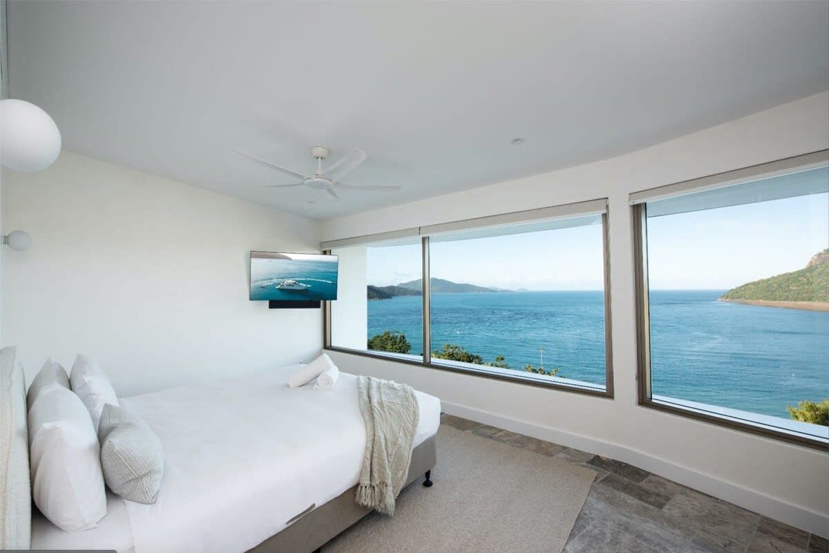 Une chambre avec un grand lit, un ventilateur de plafond et une vue sur la mer grâce à de grandes fenêtres.