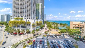 A high-rise building with a curved facade, a parking lot, and a view of the ocean. - Gym | On-site Dining | Long Stay Ideal | StayCozy (Miami)