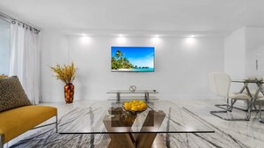 A modern living room with a glass coffee table, a sofa, a dining table with chairs, a TV on the wall, and a vase with yellow flowers. - Gym | On-site Dining | Long Stay Ideal | StayCozy (Miami)