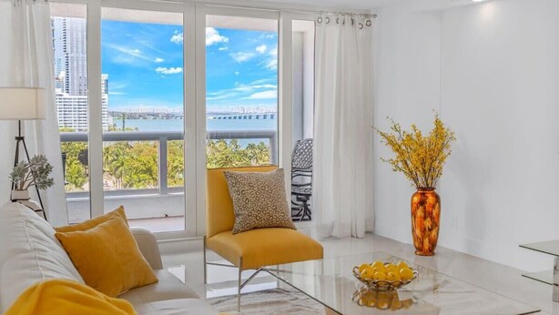 A modern living room with a sofa, armchair, and coffee table. There is a large window with a view of buildings and a bridge. - Gym | On-site Dining | Long Stay Ideal | StayCozy (Miami)