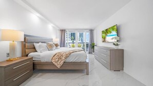 A modern bedroom with a bed, bedside tables, a dresser, and a large window with a view of buildings and a beach. - Gym | On-site Dining | Long Stay Ideal | StayCozy (Miami)