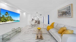 Un salon moderne avec un téléviseur à écran plat, une table basse en verre, un canapé blanc agrémenté de coussins jaunes et un tableau encadré au mur.