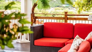 Eine Rattan-Couch mit roten Kissen auf einem Balkon, ein Glastisch und Blick ins Grüne.