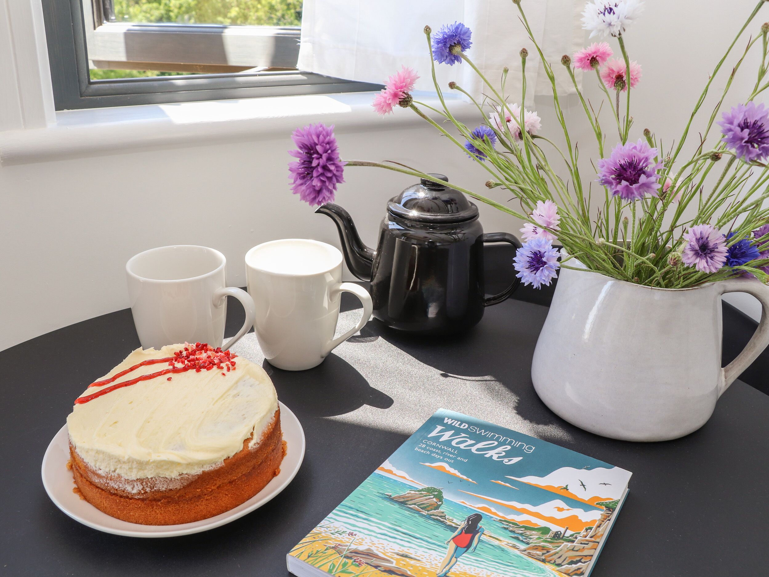 Une table avec un gâteau, deux tasses, une théière, un livre et un vase de fleurs.