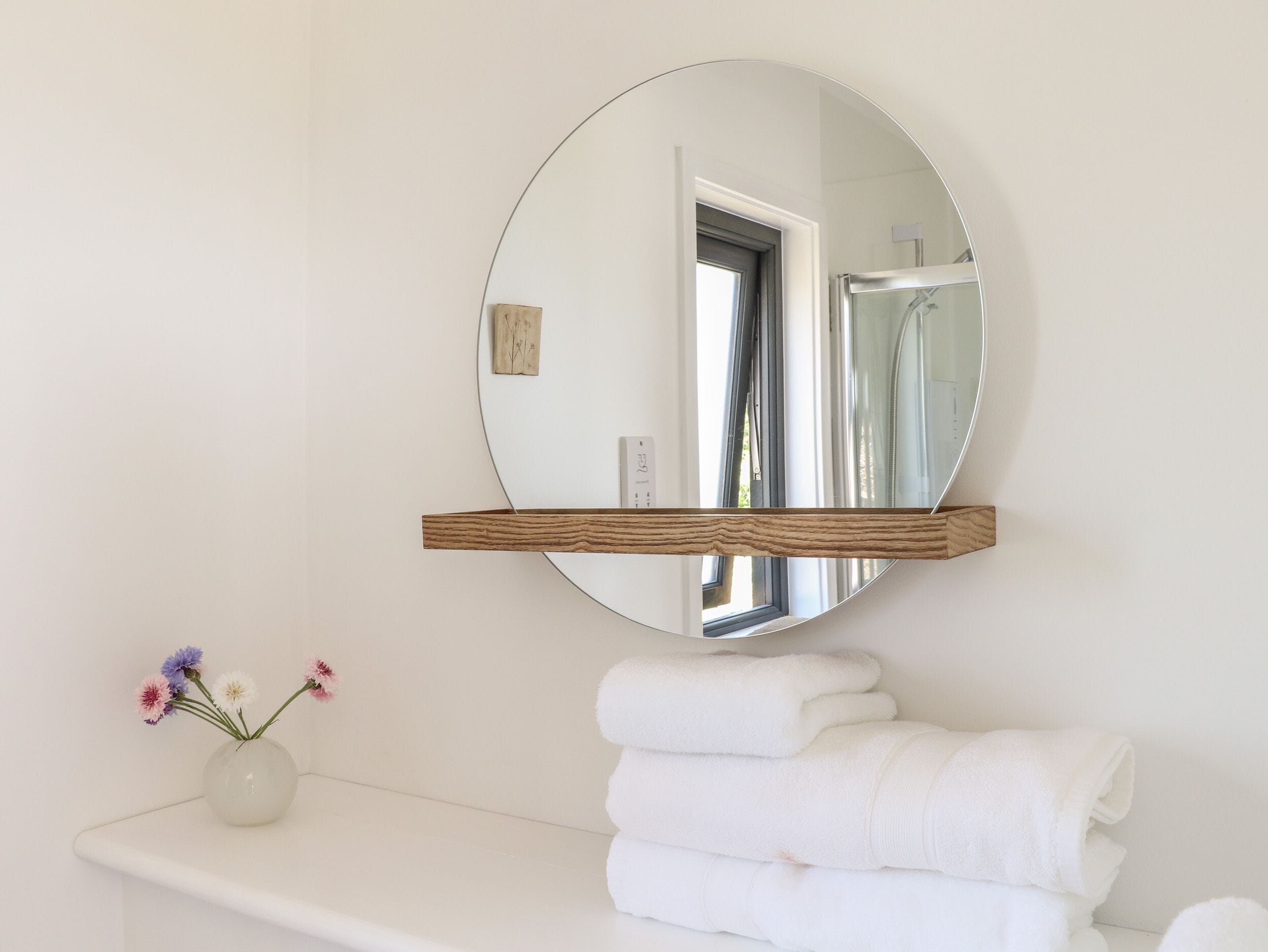 A bathroom with a round mirror, a wooden shelf, a vase with flowers, and stacked towels.