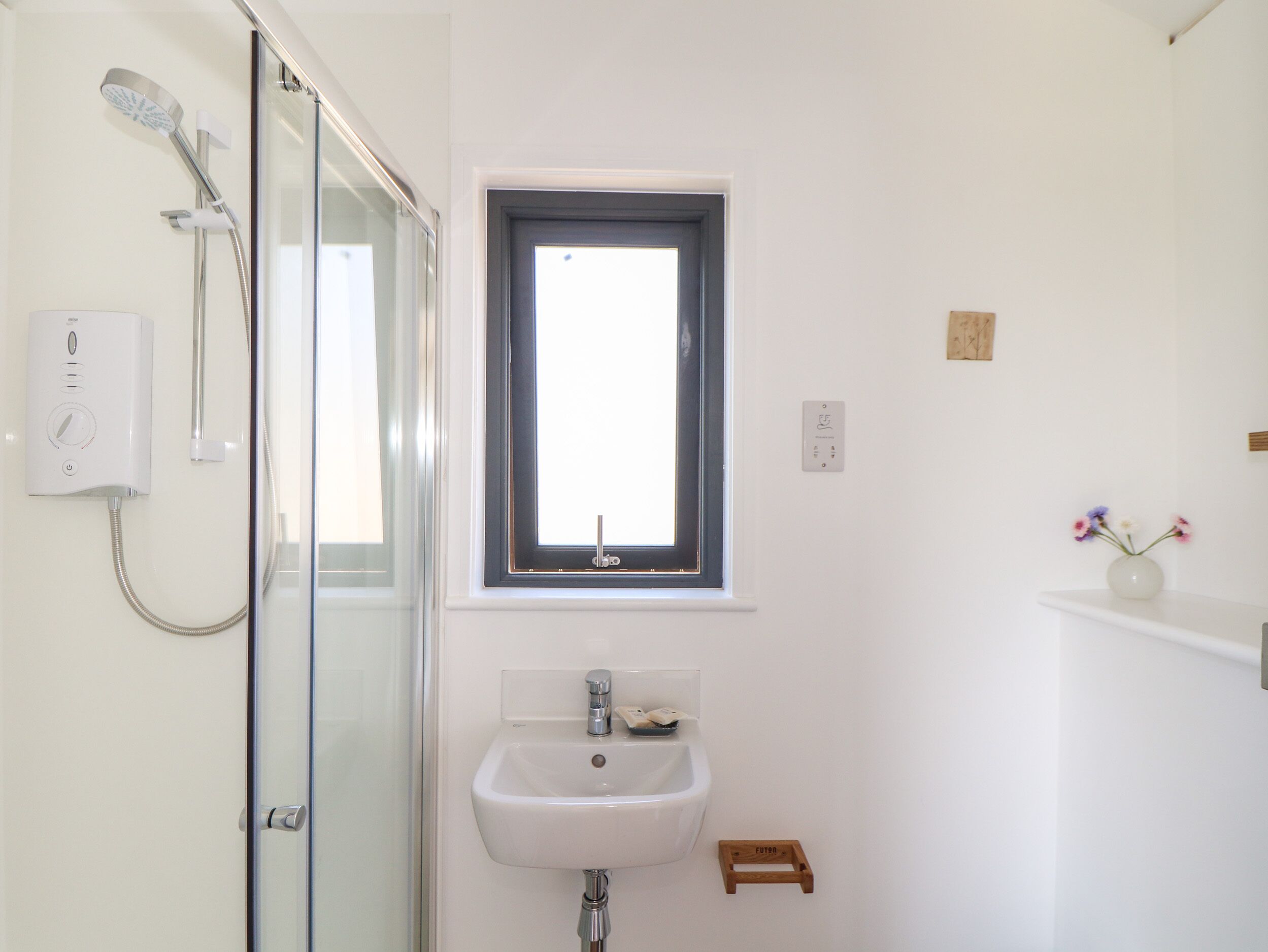 A bathroom with a glass shower enclosure, a white sink, a window, and a small wooden stool.