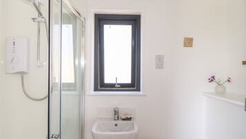 A bathroom with a glass shower enclosure, a white sink, a window, and a small wooden stool.