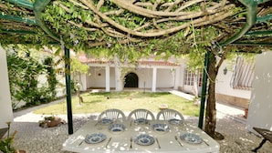 A covered outdoor dining area with a table set for six, surrounded by white chairs, under a pergola with climbing vines.