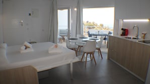 Moderni hotellihuone, jossa on sänky, ruokapöytä tuoleineen, keittiö sekä parveke, jolta on näkymä.