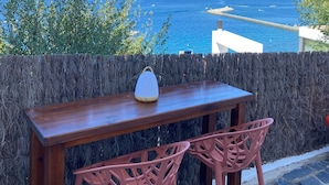Een houten tafel met twee roze stoelen en een witte lamp op een balkon met uitzicht op de kust, de zee en verre gebouwen.