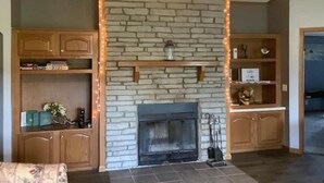 A living room with a stone fireplace, wooden shelves, and a ceiling fan.