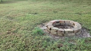 A backyard with a stone fire pit surrounded by grass, a wooden deck, and a house in the background.