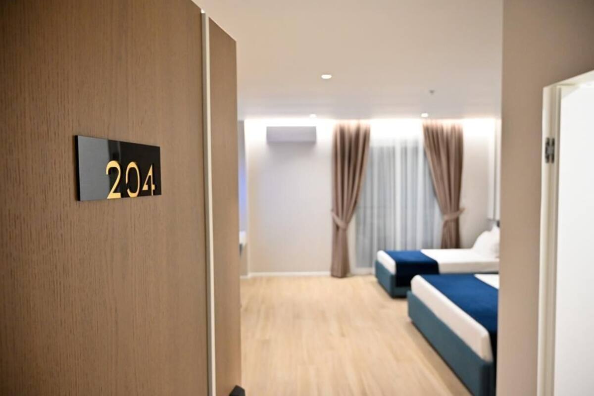 A hotel room with a wooden floor, two beds with blue bedding, a wall-mounted number sign '204', and a window with curtains.