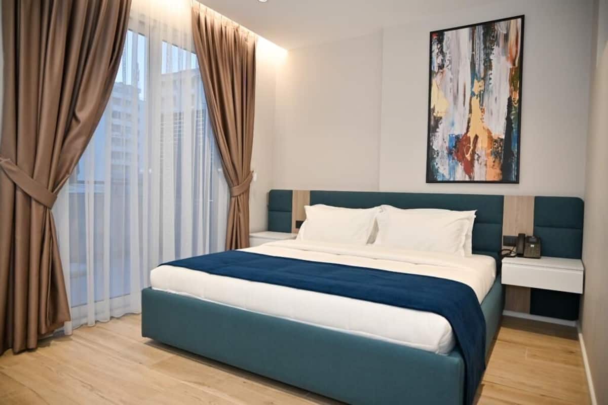 A modern hotel room with a large bed, a bedside table, a nightstand, and a painting on the wall.